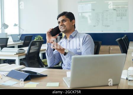 Smiling excited project manager sitting at office desk and talking on phone with client or coworker Stock Photo