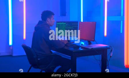 Young african man writing hacker programs in dark room with neon lights. He working on computer with two monitors. Stock Photo