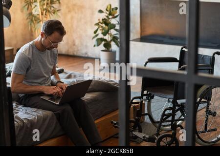 Portrait of modern disabled man using laptop while sitting on bed, wheelchair in foreground, copy space Stock Photo