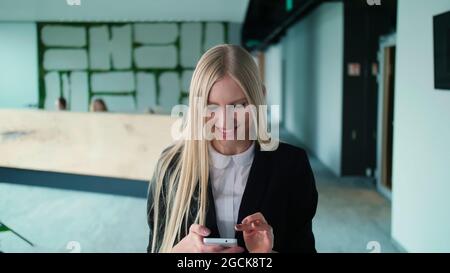 Pretty blond woman with long hair wearing elegant black suit and smiling charmingly at camera inside of modern office Stock Photo