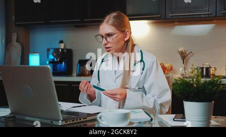 Video call to patient: female doctor in medical gown explaining how to take medicine. She using computer from home. Stock Photo
