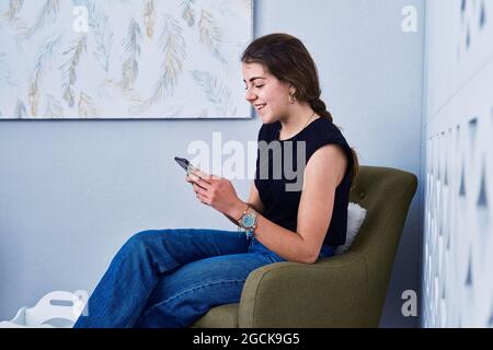 Side view of positive young female in casual outfit sitting in armchair and messaging on mobile phone Stock Photo