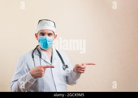 Young doctor portrait in white suit and protective breathing mask glasses and stethoscope pointing his fingers right into copyspace isolated Stock Photo