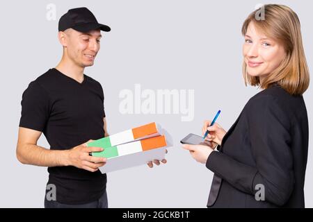 Pretty business smiling young woman looking at the camera putting signature on the tablet for the delivering service of two boxes.  Stock Photo