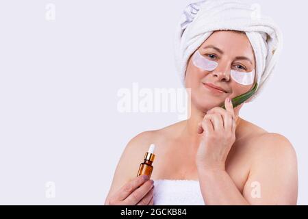 Happy smiling attractive blonde woman with the towel on the head using essential oil and aloe leaf for facial rejuvenation. Stock Photo