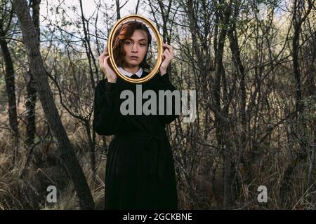 Unemotional female in black dress standing in autumn forest at Halloween and looking at camera through hole in wooden frame Stock Photo