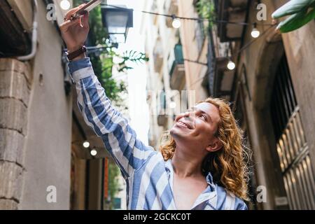 Positive handsome male with long hair taking self portrait on smartphone while standing in street in summer Stock Photo