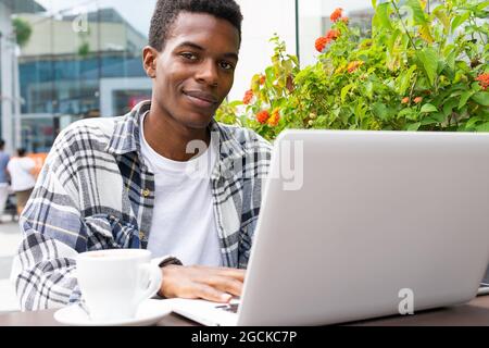 Happy African American male freelancer browsing and working remotely on laptop in outdoors cafe while sitting looking at camera at table with cup of c Stock Photo