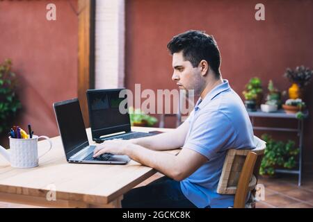 man sitting on a garden terrace using two laptops to trade in cryptocurrencies Stock Photo