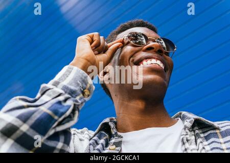Low angle of handsome smiling African American male wearing trendy round sunglasses looking away on bright blue background in street in summer Stock Photo