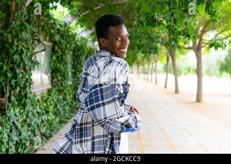 Back view of glad African American male in plaid shirt standing in city street and looking at camera over shoulder Stock Photo