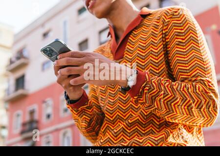 From below cropped unrecognizable side view of young Hispanic guy with Afro hair in stylish colorful outfit browsing mobile phone while standing on ra Stock Photo