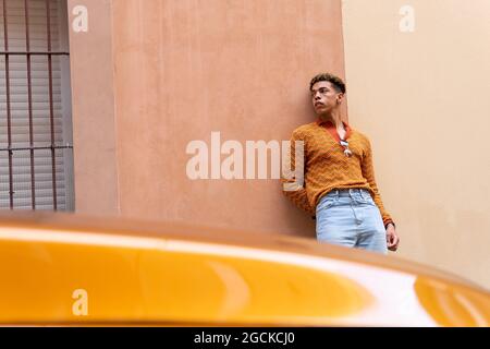 Young stylish ethnic curly haired guy in trendy outfit leaning against wall near parked modern orange automobile on urban street Stock Photo