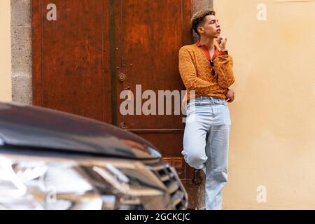 Young stylish ethnic curly haired guy in trendy outfit leaning against wall near parked modern automobile on urban street Stock Photo