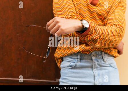 Crop anonymous male in stylish colorful striped shirt and jeans with wristwatch and sunglasses standing near wall Stock Photo