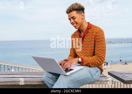Positive young ethnic male student in stylish outfit typing on laptop keyboard while sitting on bench near sea on urban beach Stock Photo