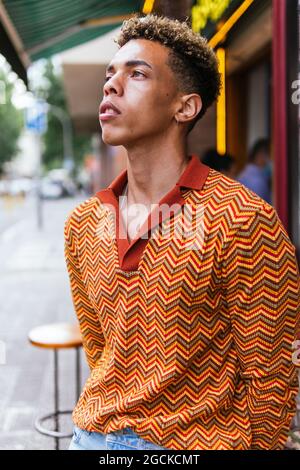 Young ethnic curly haired guy in stylish colorful striped shirt standing on the street looking away pensively Stock Photo