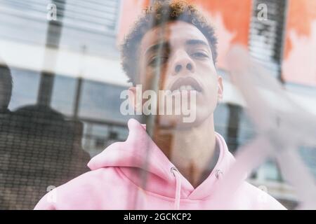 Through glass of serious young ethnic hipster guy with Afro hairstyle dressed in pink hoodie at window and looking at camera Stock Photo