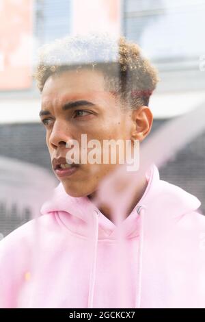 Through glass of serious young ethnic hipster guy with Afro hairstyle dressed in pink hoodie at window and looking away Stock Photo