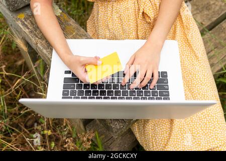 From above of crop anonymous female buyer making purchase with plastic card during online shopping via netbook while sitting on bench in park Stock Photo