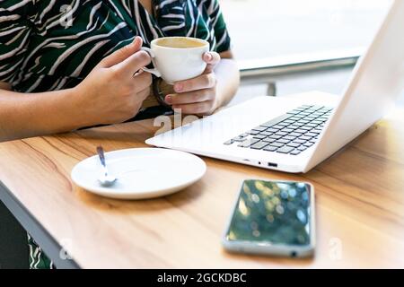 Crop anonymous female freelancer drinking coffee and working with laptop while sitting at table in cafe Stock Photo