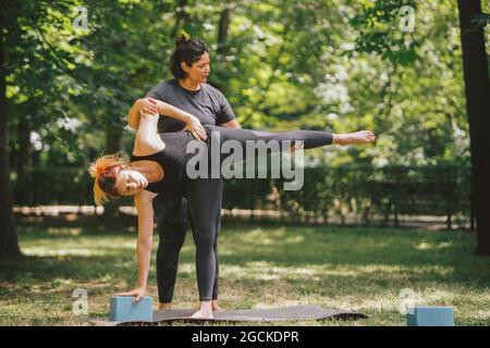 Yoga teacher supporting female in sportswear performing Ardha Chandrasana pose on mat during class in sunny park Stock Photo