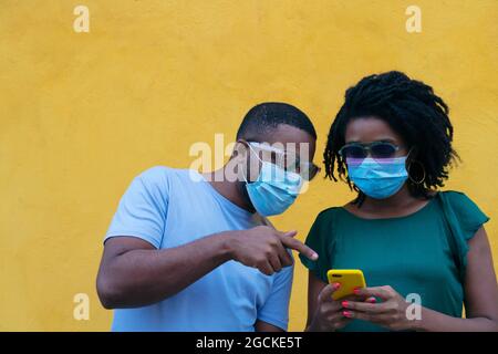 Black couple with mask using a mobile phone leaning against a yellow wall Stock Photo