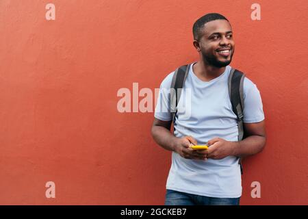 African man with backpack and mobile phone smiling while leaning on wall Stock Photo
