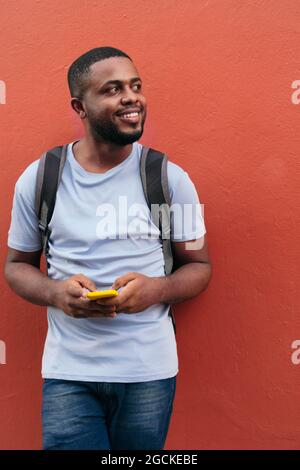 Black man with backpack and mobile phone smiling while leaning on wall Stock Photo