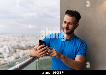 Young focused Hispanic male with dark hair and beard in t shirt standing on balcony of multistory building and using mobile phone Stock Photo