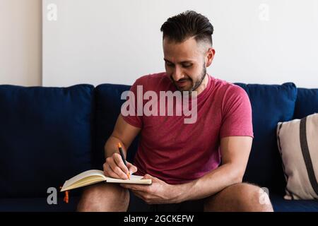 Cheerful latin man writing on notebook while sitting on sofa at home Stock Photo
