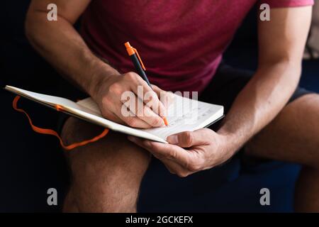Crop of anonymous latin man writing on notebook while sitting on sofa at home Stock Photo