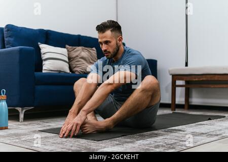 Concentrated athletic male doing stretch exercises near sofa while training at home and looking away Stock Photo