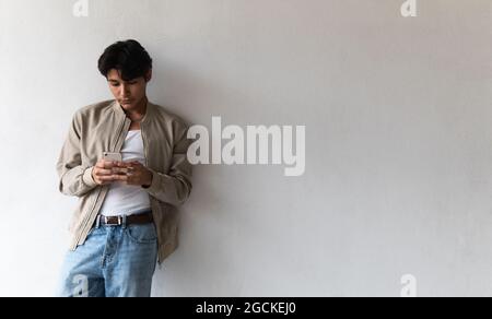 Young focused latin man in casual clothes using phone while leaning on white wall Stock Photo