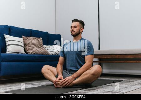Concentrated athletic male doing stretch exercises near sofa while training at home and looking away Stock Photo