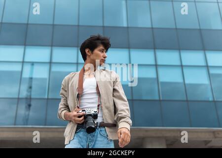 From below young latin man photographer holding photo camera looking away against modern building Stock Photo