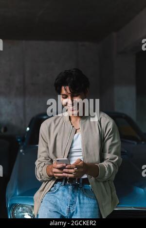 Young smiling latin man in casual clothes using phone while leaning on vintage car in parking lot Stock Photo