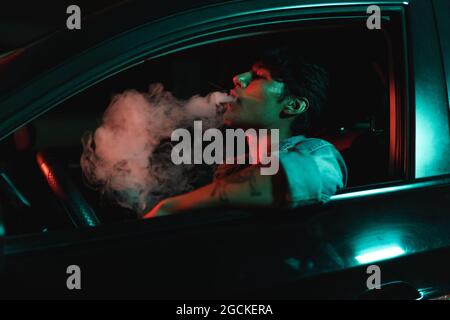 Young latin man sitting in car and smoking under color lights at night Stock Photo