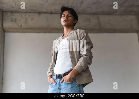 From below portrait of young latin man looking confidently at camera with hands on pockets against neutral background Stock Photo