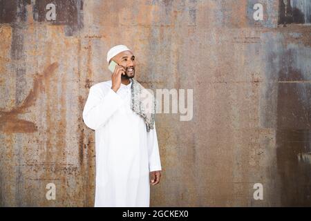 Cheerful Muslim male in traditional clothes smiling and talking on the cellphone while standing near shabby wall on street Stock Photo