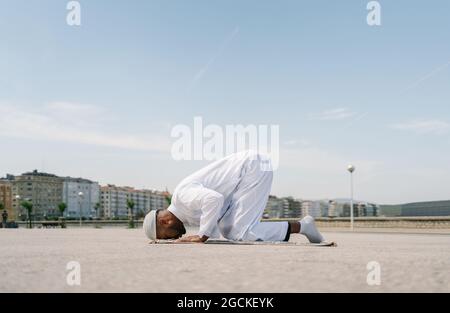 Muslim male kneeling on rug and touching ground with forehead while praying on sandy beach on sunny day Stock Photo