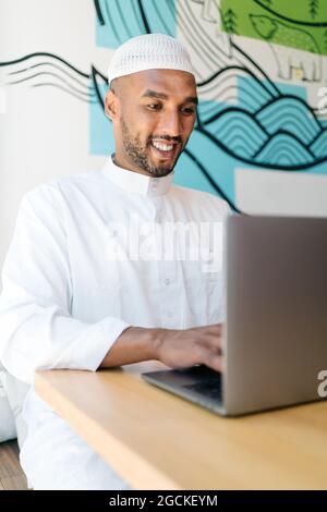 Happy Muslim male in authentic clothes sitting at table and browsing netbook in cafeteria Stock Photo