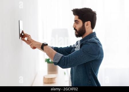 Automation domestic system. Arab man using digital tablet on wall with smart home control system app, adjusting house settings, standing in light livi Stock Photo