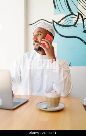 Happy Muslim male in authentic clothes sitting at table and browsing netbook in cafeteria Stock Photo