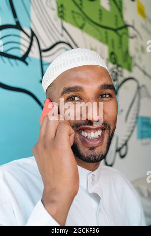 Cheerful Muslim male in traditional clothes smiling and using cellphone while standing near wall on street Stock Photo