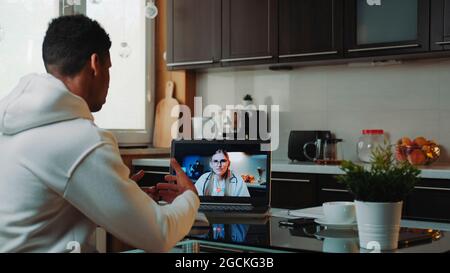Black man making video consultation with female doctor on computer. He explaining disease symptoms while sitting in the kitchen at home. Stock Photo