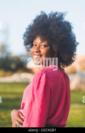 Side view of charming African American female with curly hair smiling at camera while standing on sunny day in park Stock Photo