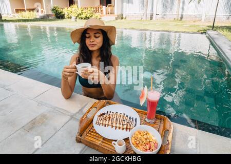 Cheerful female tourist leaning on poolside while drinking coffee against tray with yummy breakfast in sunlight Stock Photo