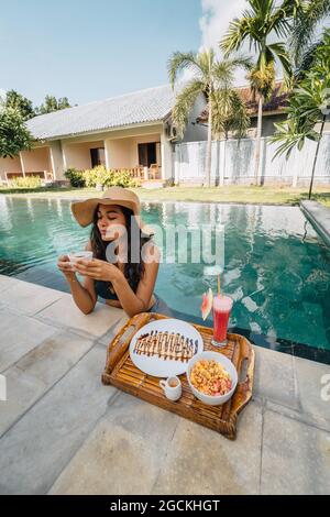 Cheerful female tourist leaning on poolside while drinking coffee against tray with yummy breakfast in sunlight Stock Photo