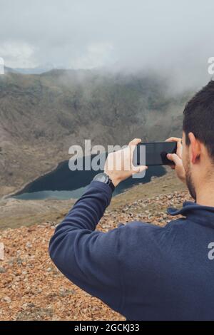 Back view of unrecognizable male hiker standing on hill and taking picture of amazing highlands on smartphone on misty day during vacation in Wales Stock Photo